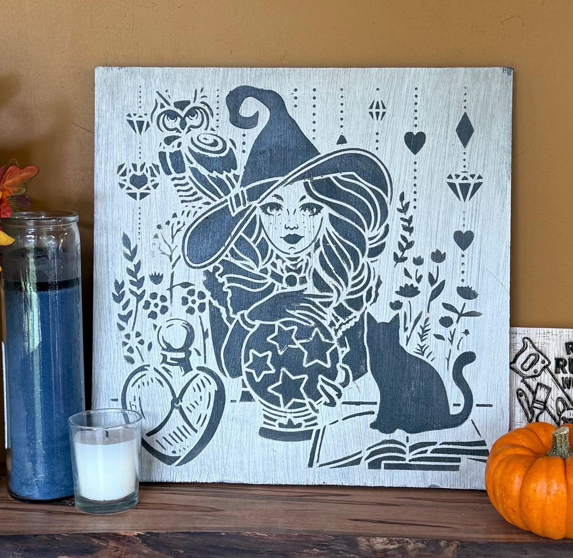 Decorative wall art with a witch design on a wooden surface with a candle and pumpkin.
