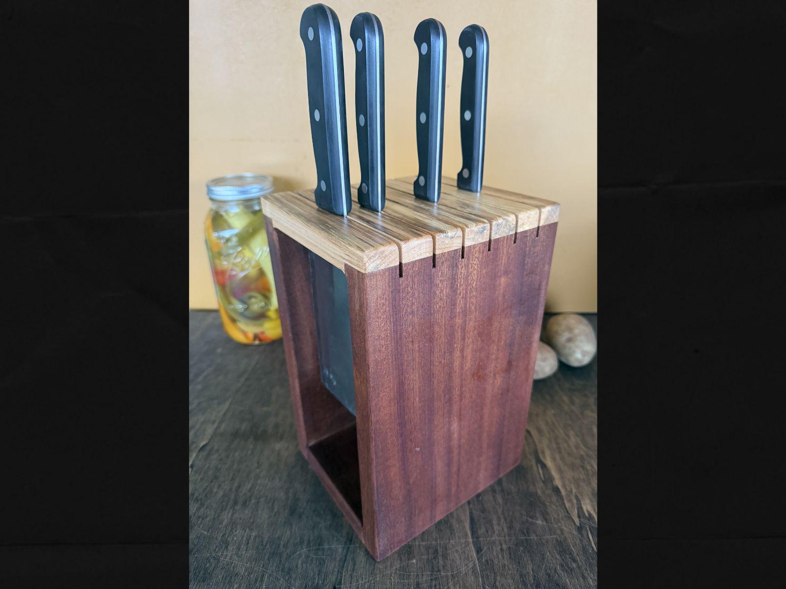 Set of knives in a wooden block on a wooden surface with a jar of pickles in the background.