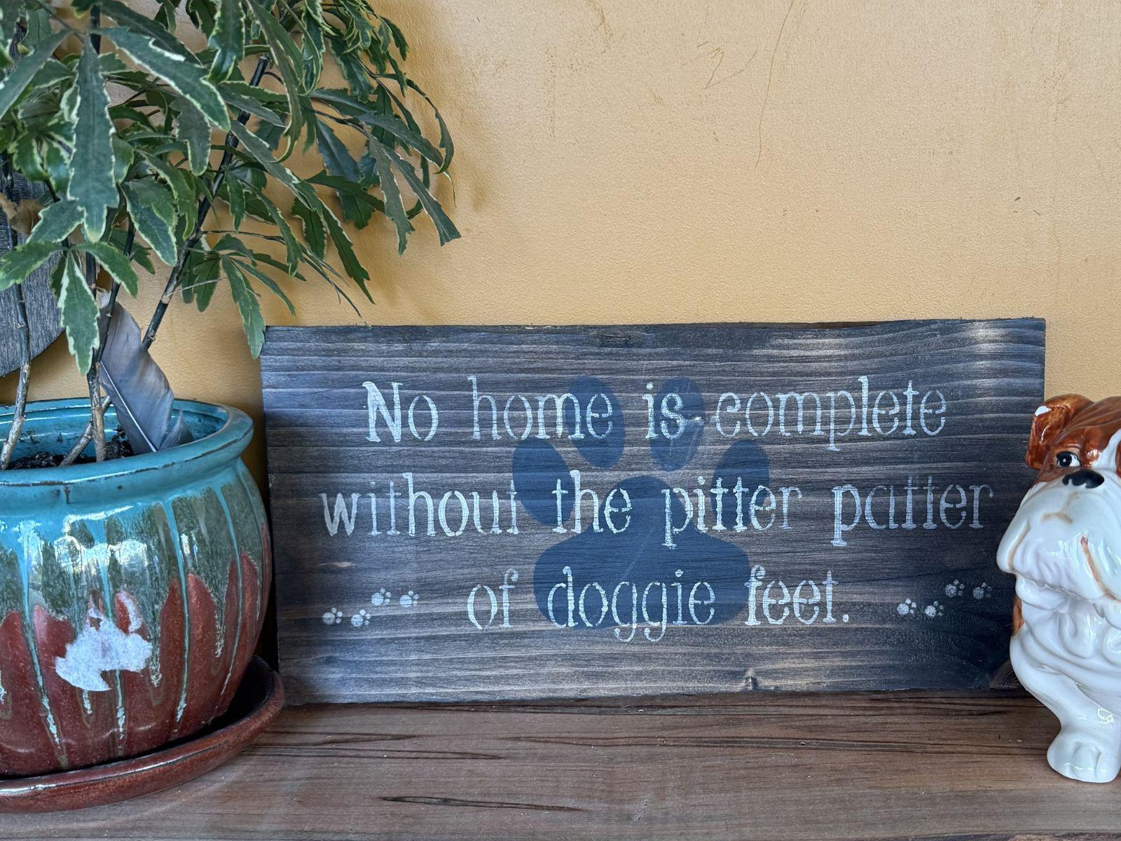 Decorative sign that says "No home is complete without the pitter patter of doggie feet" next to a potted plant and a small dog figurine on a wooden surface.
