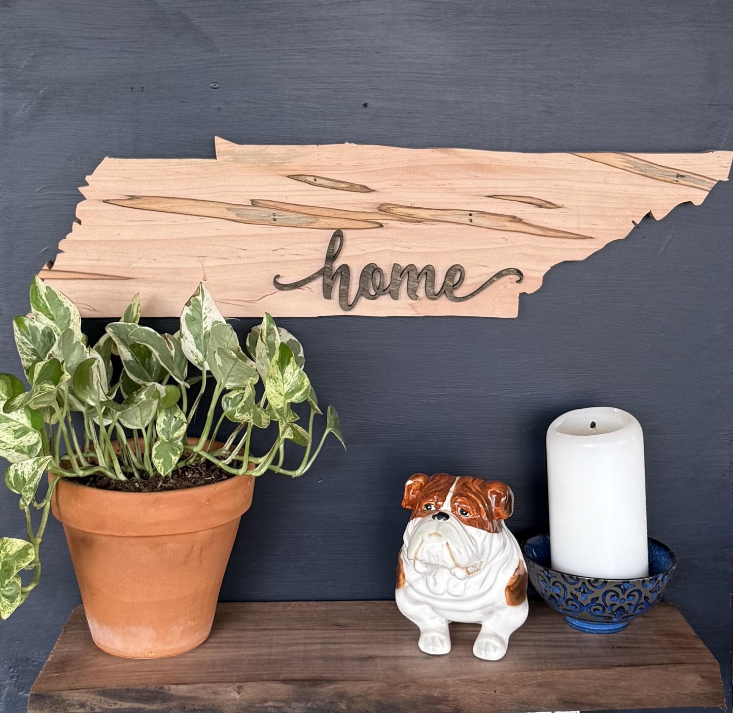 Wood Tennessee shaped sign with 'home' engraved, potted plant, ceramic dog figurine, and candle on a dark surface.