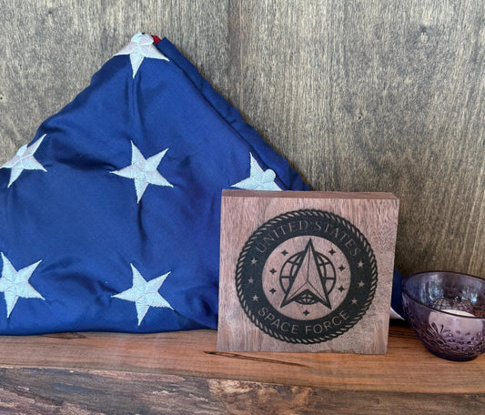 Wooden plaque with 'United States Space Force' design next to a folded American flag on a wooden surface.