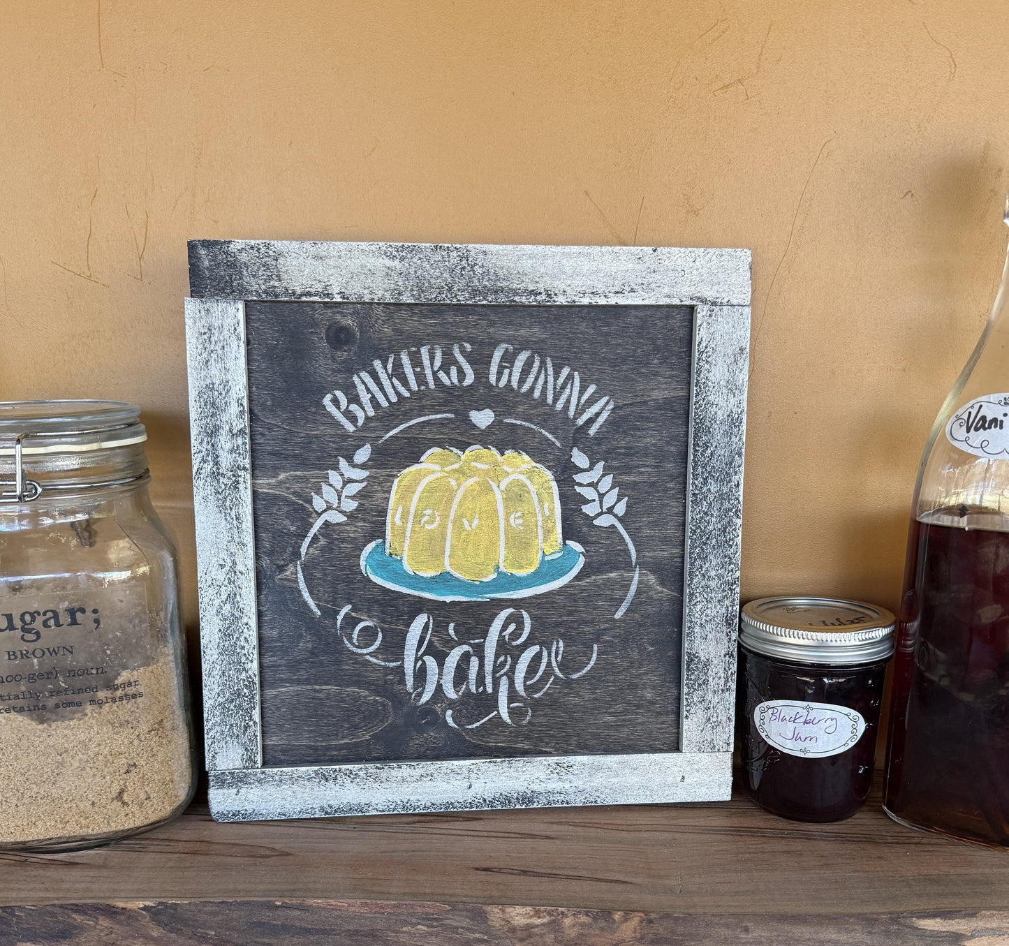 Wooden sign with a yellow cake illustration and text that says "Bakers Gonna Bake" on a shelf with jars and bottles.