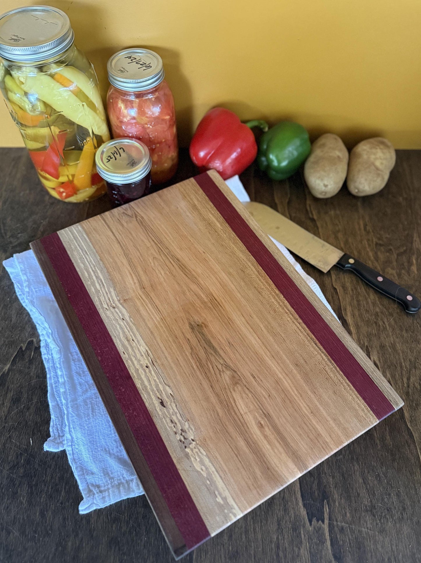 Wooden cutting board on a wooden surface with jars, vegetables, and a knife.