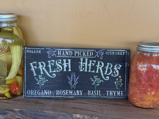 Wooden sign with 'Fresh Herbs' text between two jars on a wooden surface.