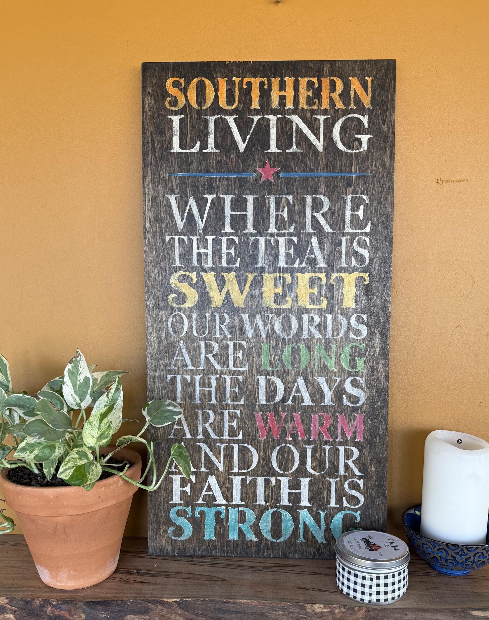 Decorative sign with text “Southern Living – Where the Tea Is Sweet, Our Words Are Long, The Days Are Warm, and Our Faith Is Strong.” on a wooden surface with a plant and candle in the background.