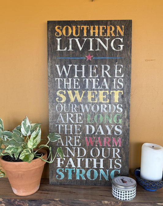 Decorative sign with text “Southern Living – Where the Tea Is Sweet, Our Words Are Long, The Days Are Warm, and Our Faith Is Strong.” on a wooden surface with a plant and candle in the background.