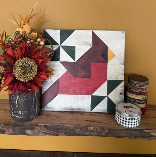 Geometric barn quilt with an abstract Cardinal bird motif on a wooden shelf with a plant and candles.