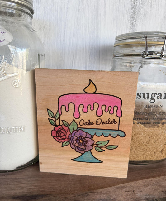 Wooden sign with a colorful cake illustration and 'Cake Dealer' text, placed between two glass jars.