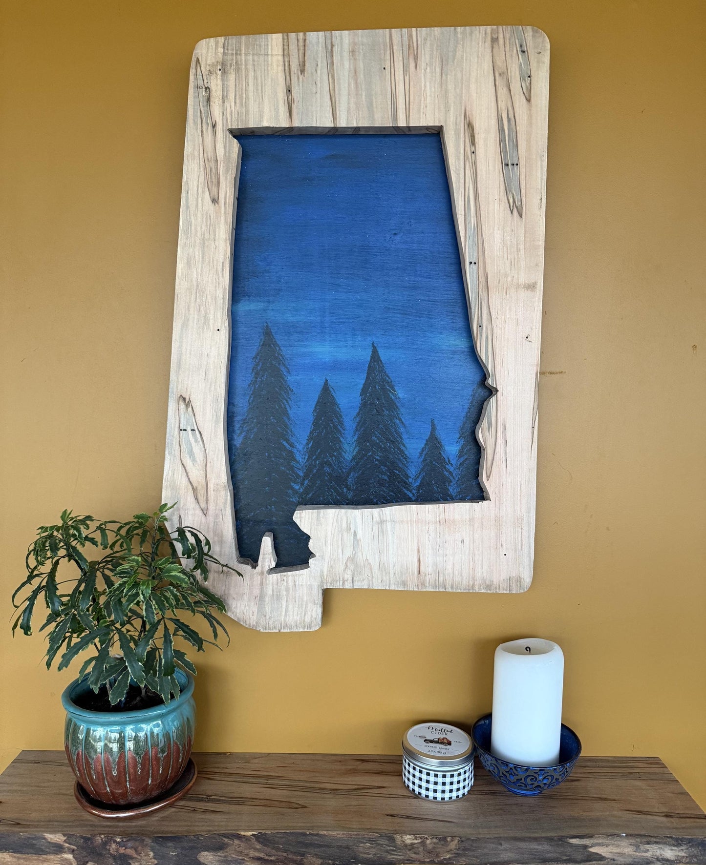 Wood cutout of the State of Alabama with hand painted night sky hanging on a yellow wall.
