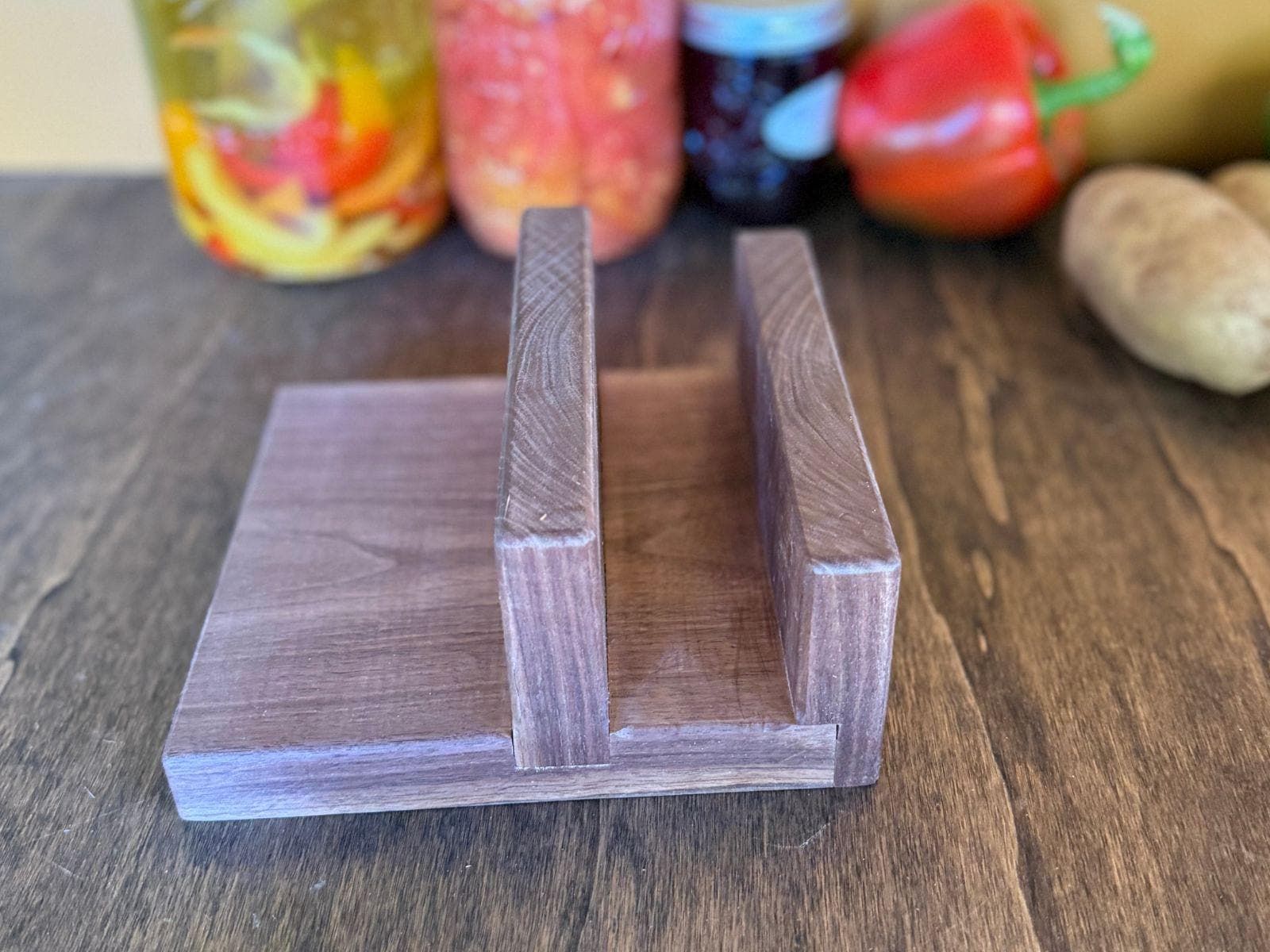 Wooden cutting board holder on a wooden surface with jars and vegetables in the background.